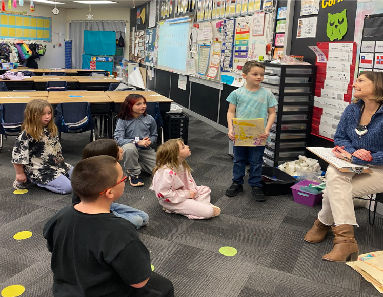 Students gathered on the carpet intently listening to the student presenting his tornado project