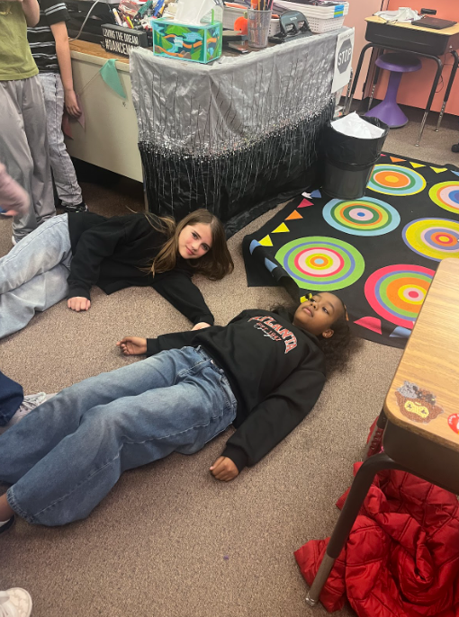 Two students laying on the carpet in their classroom to make a letter