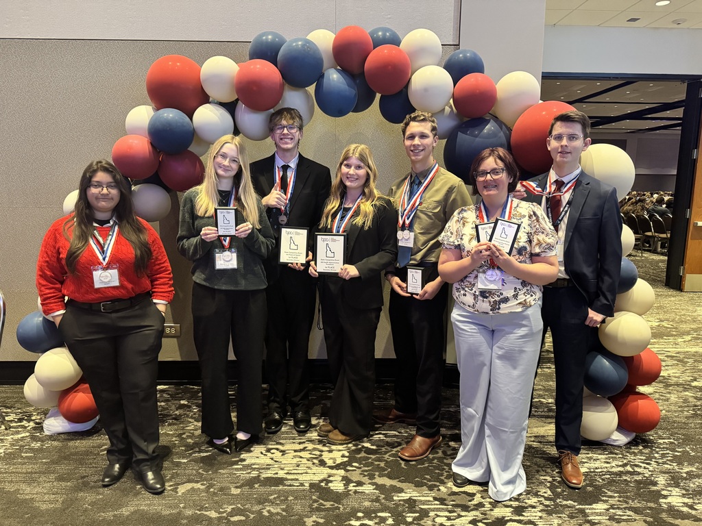 Seven students pictures holding various plaques in front of a balloon arch in a conference room