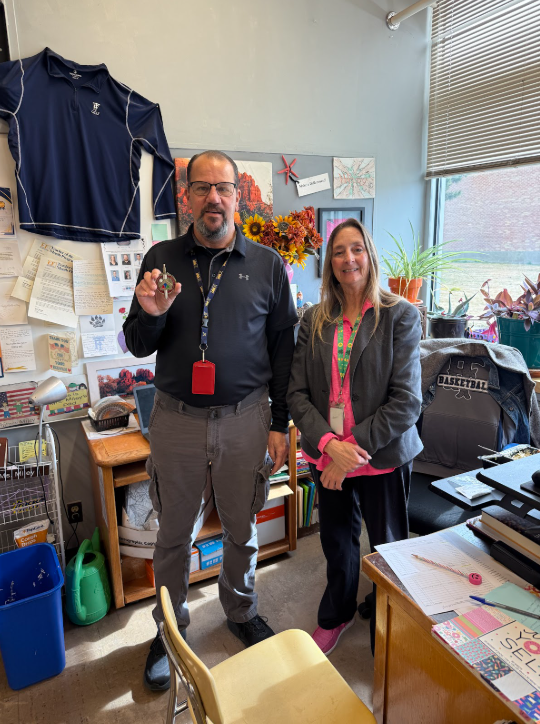 Two staff member standing together in an office, one holding up a army coin.