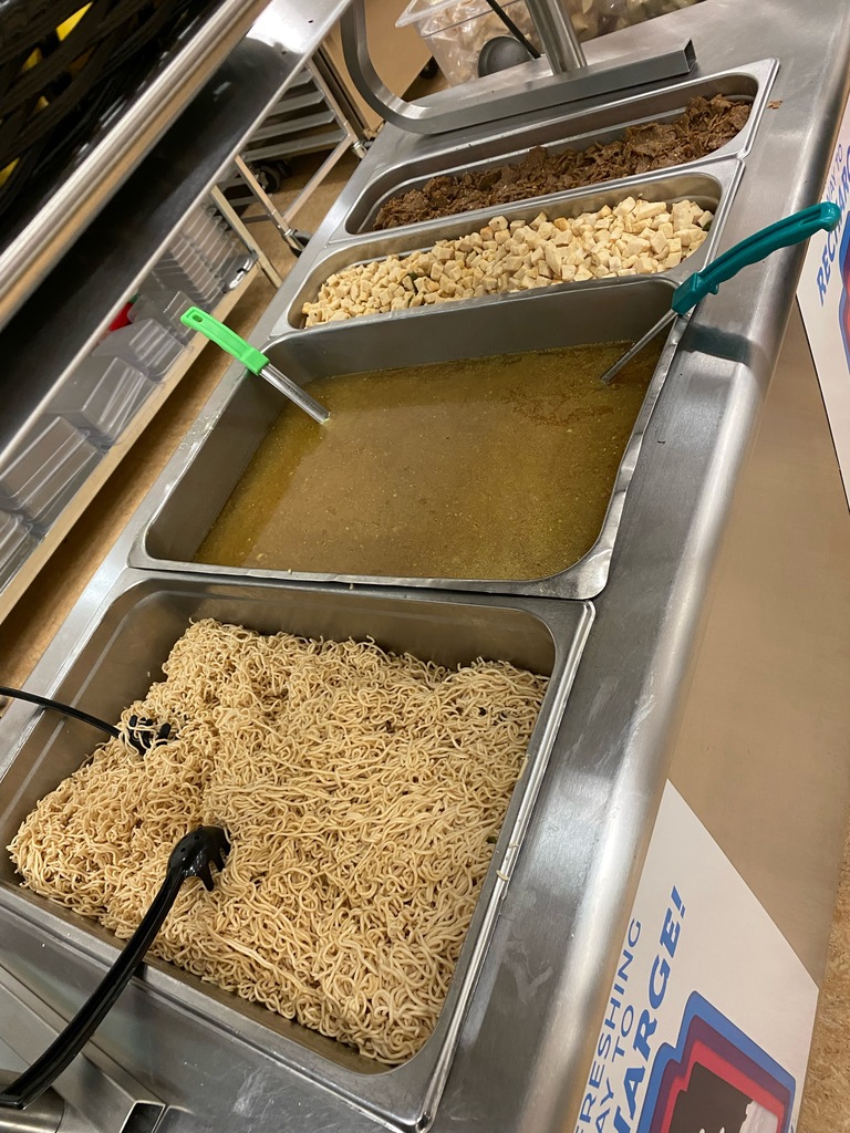 a picture of a cafeteria serving table with metal trays filled with ramen noodles , broth, and meat