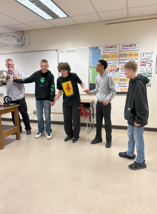 Three students holding hands, the last one touching a metal ball while the teacher laughs at them