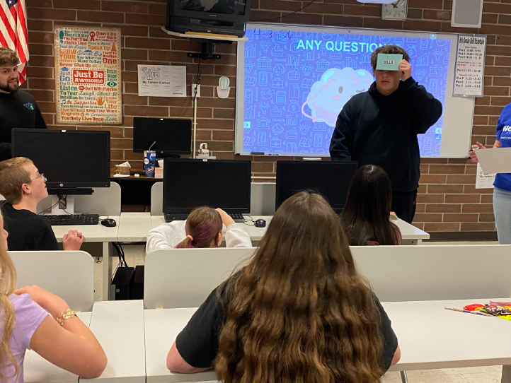 A student standing in front of the class holding a blue card to his forehead with a word on it to play a game to test the kids about what they learned.