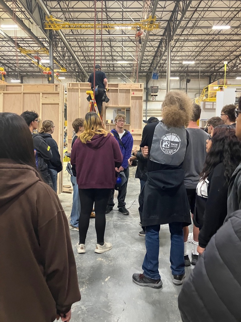 Students gathered around their guide who is explaining the wall of a camping trailer being built behind him.
