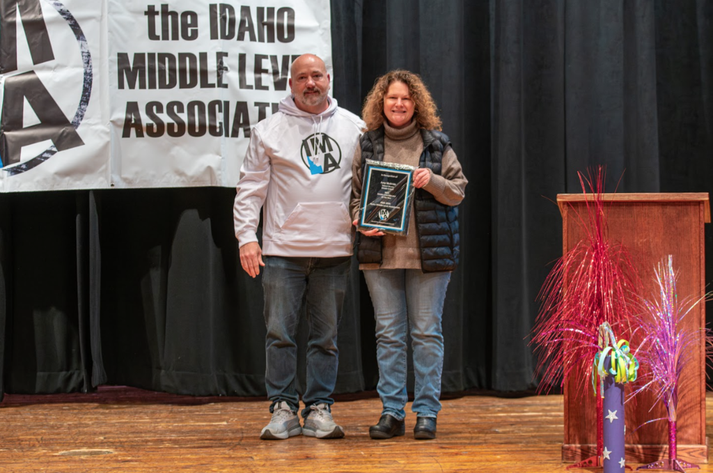 A male and a female standing on a stage with a navy curtain behind them. The female is holding a plaque award.