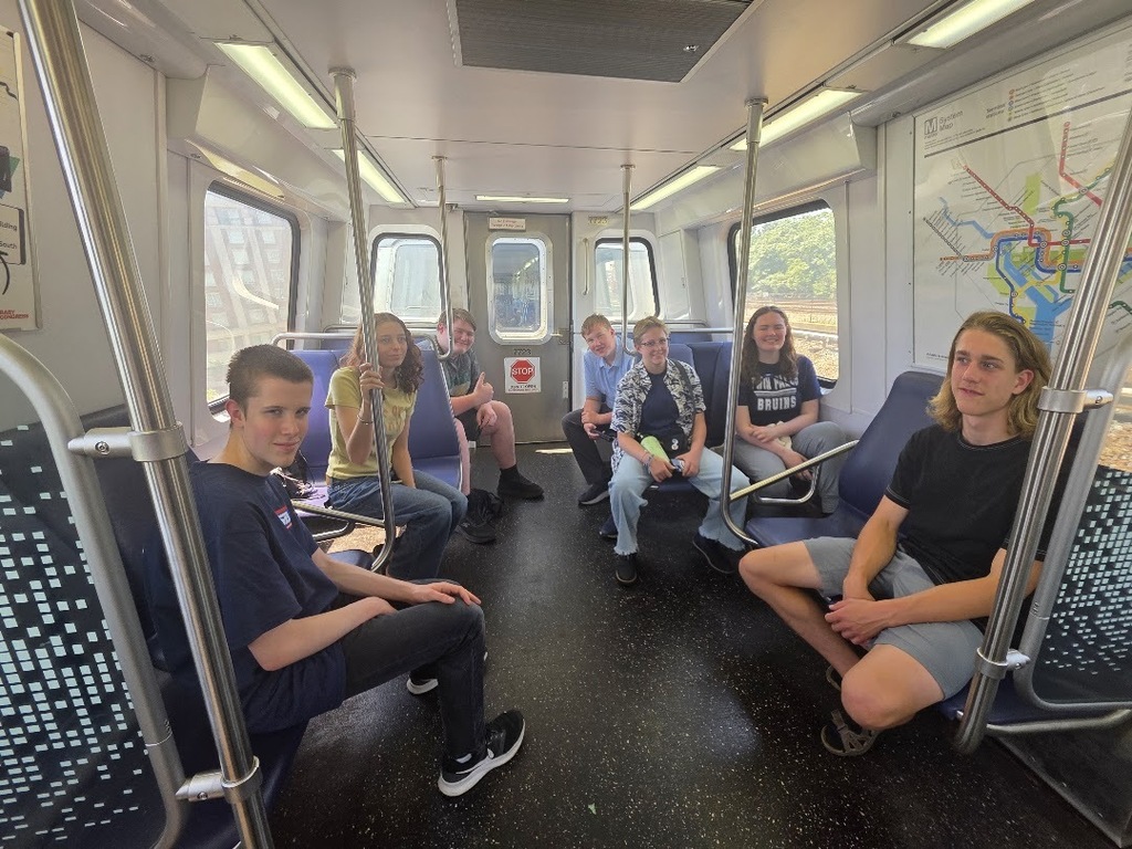Eight people in casual clothing sitting on a subway train, looking around and at the camera.