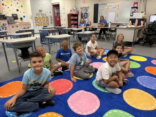 Classroom with colorful floor mats; children sit on the floor, smiling, and a teacher is seated at a desk.