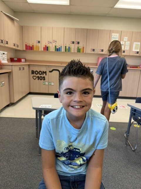 A smiling boy in a blue shirt sits at a table. Behind him, another child walks with a bag.