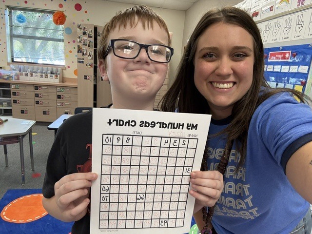 A boy in glasses and a woman in a blue shirt hold a crossword puzzle together in a classroom.