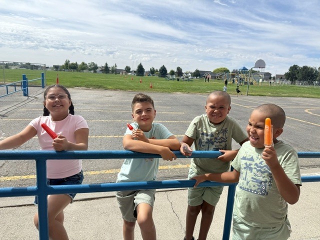 Four children in a playground, holding popsicles, standing on a blue railing. An open field is in the background.
