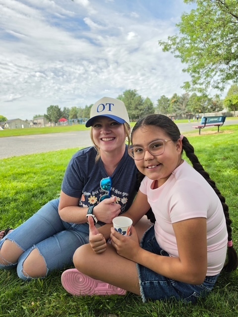 Two people sitting on grass, one wearing a cap and holding a cup, and another wearing glasses and giving a thumbs-up.