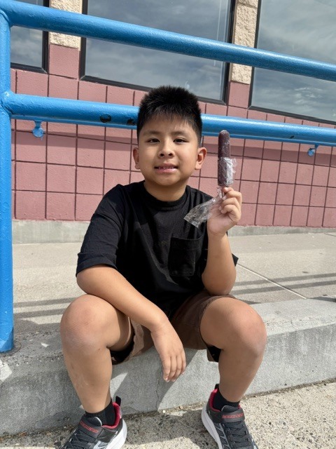 A boy sits on a concrete step, holding an ice cream bar. He wears a black t-shirt, brown shorts, and sneakers.