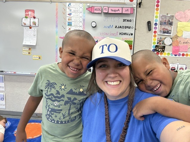 Two smiling children and a woman wearing a blue shirt with "TO" on a cap pose in a classroom.