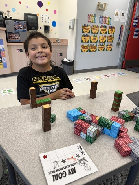 Child sitting at a table with colorful blocks, smiling, and a grid paper on the table.