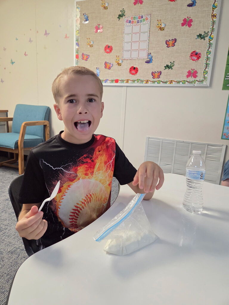 A child holds a plastic bag with a spoon while seated at a table. A water bottle and a blue chair are nearby.