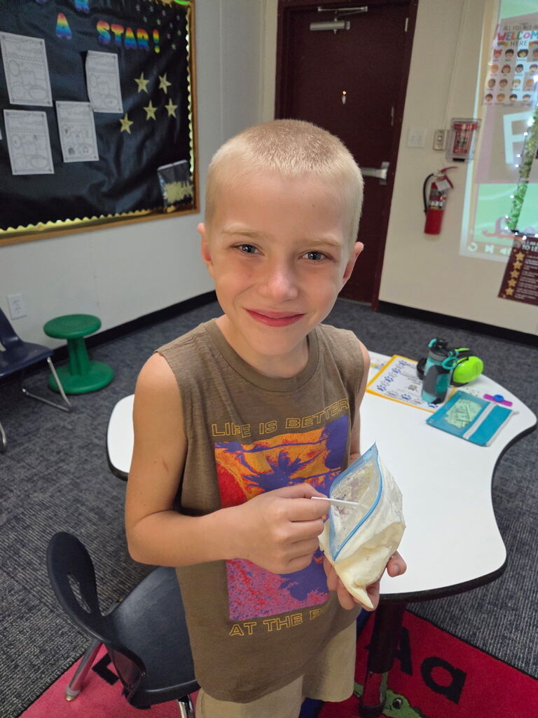 A boy holds a bag in a classroom with desks, a bulletin board, and a door.