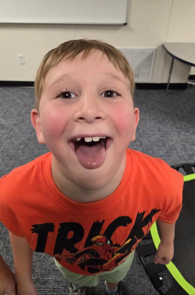A child in a room, wearing a bright orange T-shirt, smiles widely, showing teeth, and sticking out his tongue.