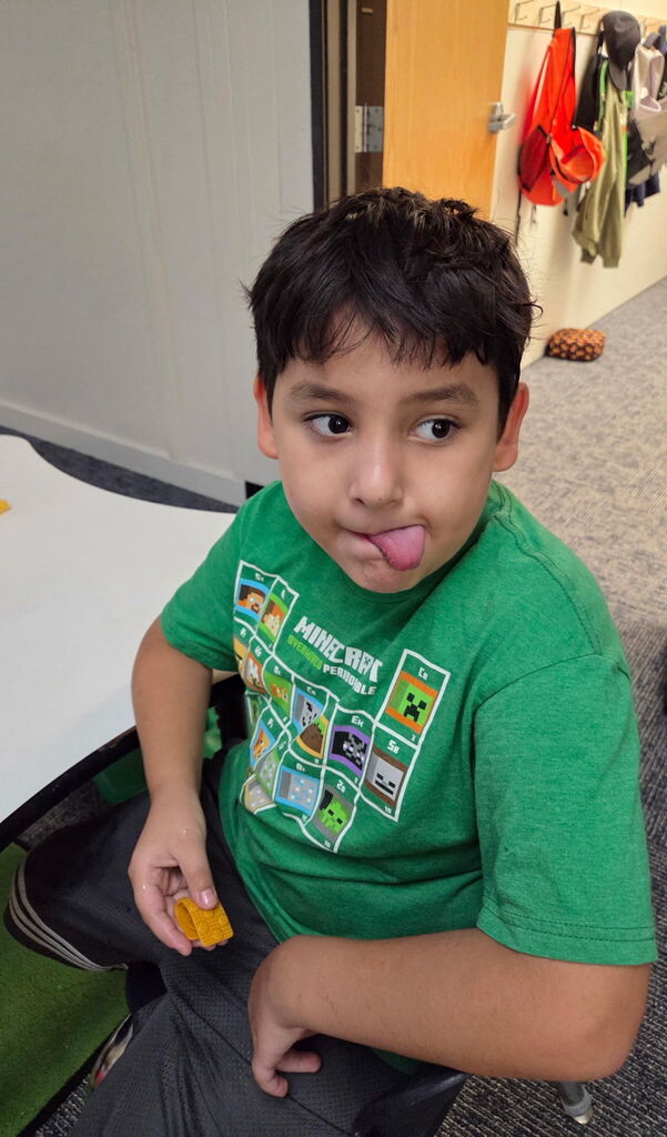 A child in a green t-shirt sticks out his tongue while sitting in a classroom.