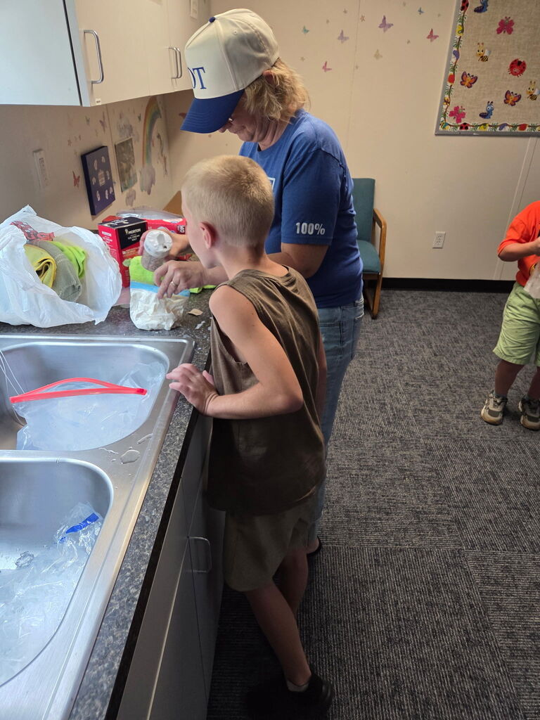 Adult in blue shirt assists a boy in brown shirt at the kitchen sink. Nearby, a child in orange shorts stands by.
