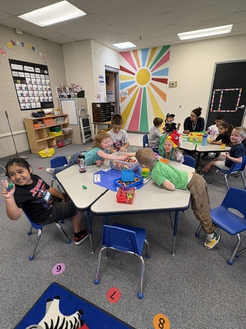 Children seated at tables, engaged in play activities with toys. Colorful sun artwork on the wall.