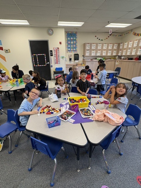 Classroom with children seated at tables. Some are playing with Legos. Tables have various items on them.
