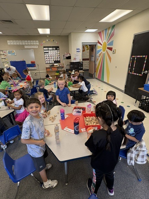 Group of children seated at tables in a classroom. Colorful decorations on the walls. One child has a snack.