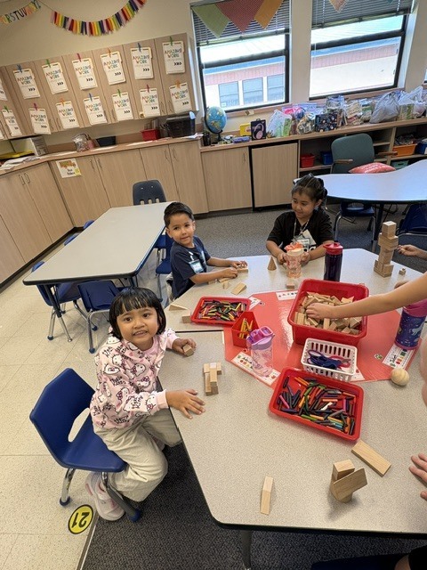 Four children sit around a table, engaged in a creative activity with blocks and art materials.