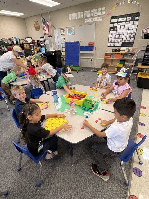 In a classroom, children sit around a table, engaged in an activity with toys and objects. Bright lighting and colorful decorations adorn the space.