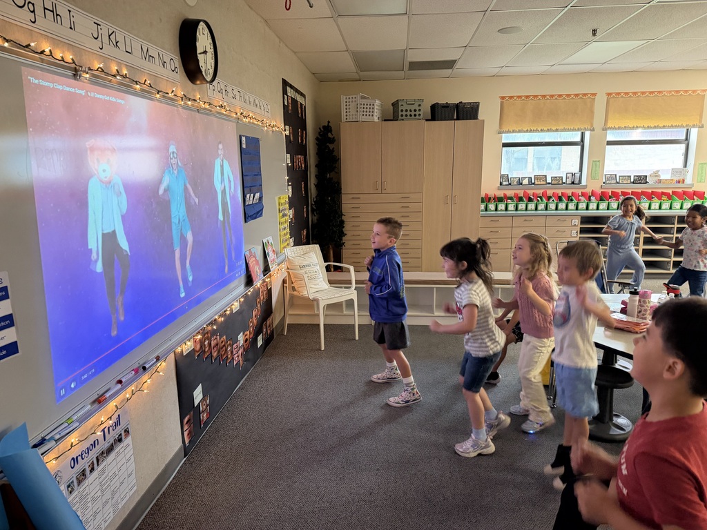 Kids in a classroom are watching a screen while some are dancing. A clock and bulletin board are on the wall.