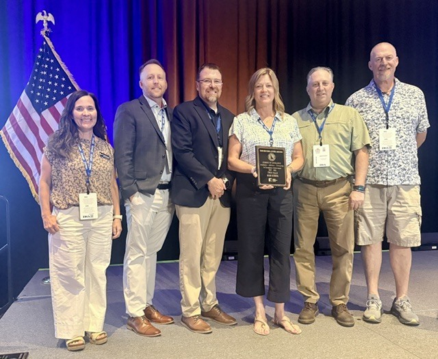 Group of educators beam brightly at the camera as they hold their award in the front of a stage.