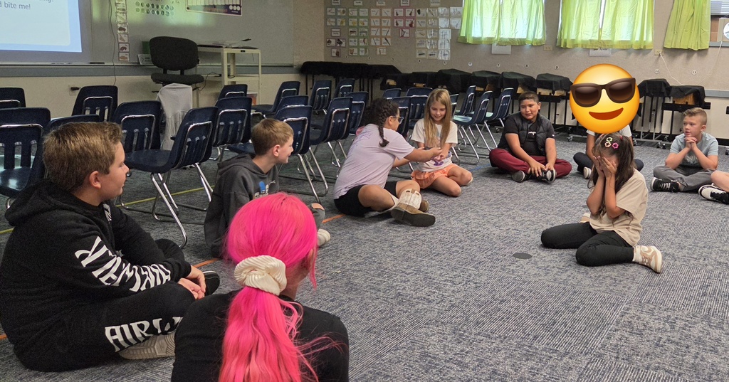 A classroom with students sitting on the floor, some in chairs. One wears sunglasses, and others have pink hair.