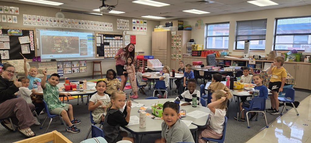 A classroom with children sitting at tables, one child standing. A projector screen is mounted on a wall.