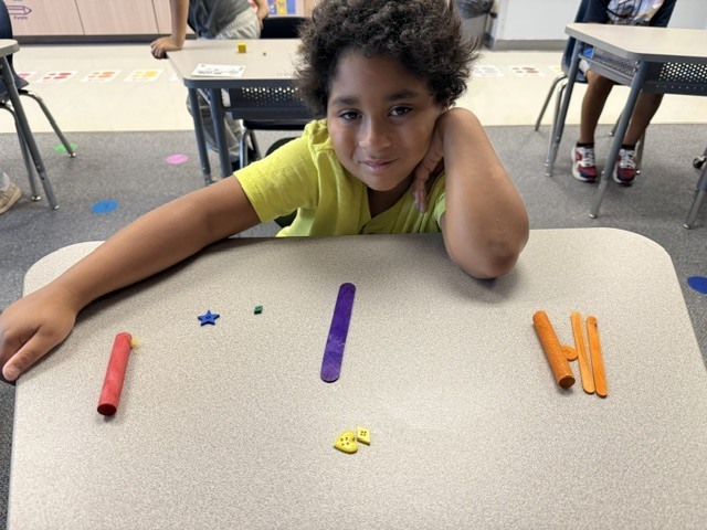 A child sits at a desk in a classroom, leaning on the table, surrounded by colorful sticks.
