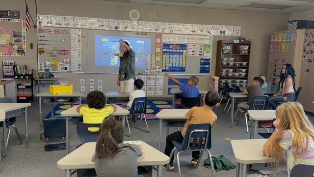Classroom with students seated at desks, a teacher stands in front of a projector screen. Walls have posters and information.