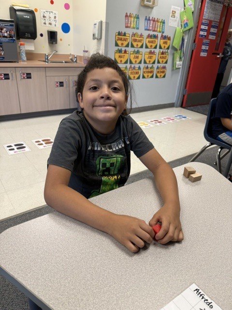 A young child sits at a table with blocks in a classroom. Behind him, there is a bulletin board.