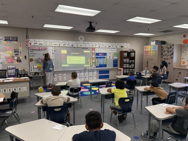A classroom with students seated at desks. A teacher stands in front of a projector screen.