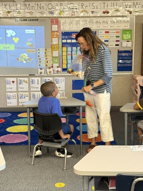 Teacher helping a student with a bag of items at a table in a classroom with colorful carpets.