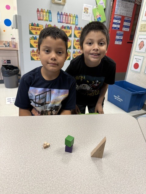 Two boys at a table, smiling, with wooden blocks in front of them. Behind them are colorful posters and a blue bin.