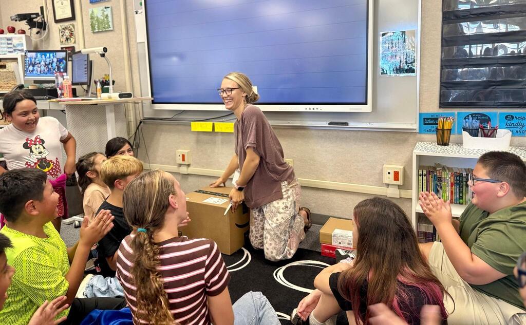 A woman stands before a classroom with a large screen on the wall, teaching a group of seated children.