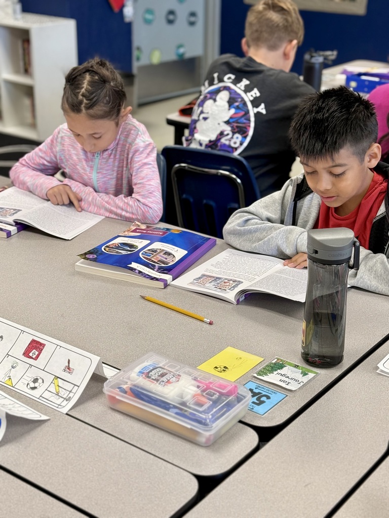 Children seated in a classroom, reading books. A girl in pink, a boy in gray, and a boy in black. Open books, a pencil, and a bottle on the table.