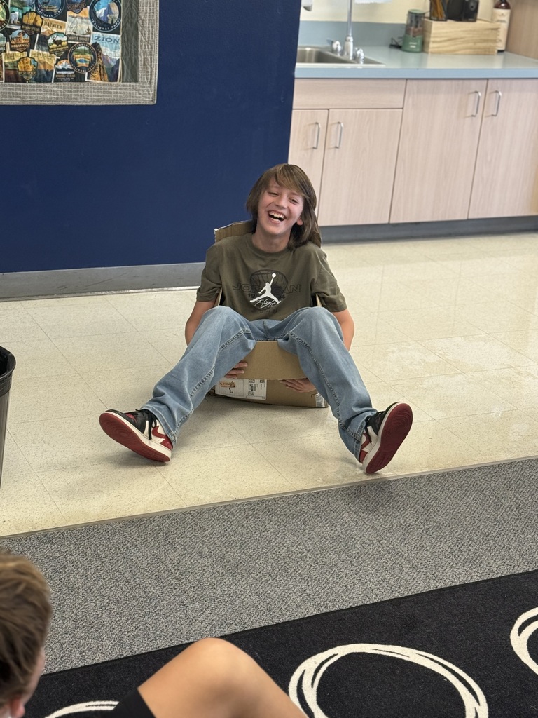 A young person in a green t-shirt and jeans sits on a box in a room with a blue wall and a kitchen counter.