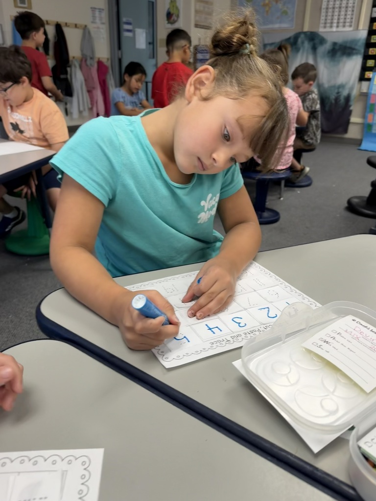 A child in a light blue shirt works on a worksheet, seated at a table. Other children are seated behind her.