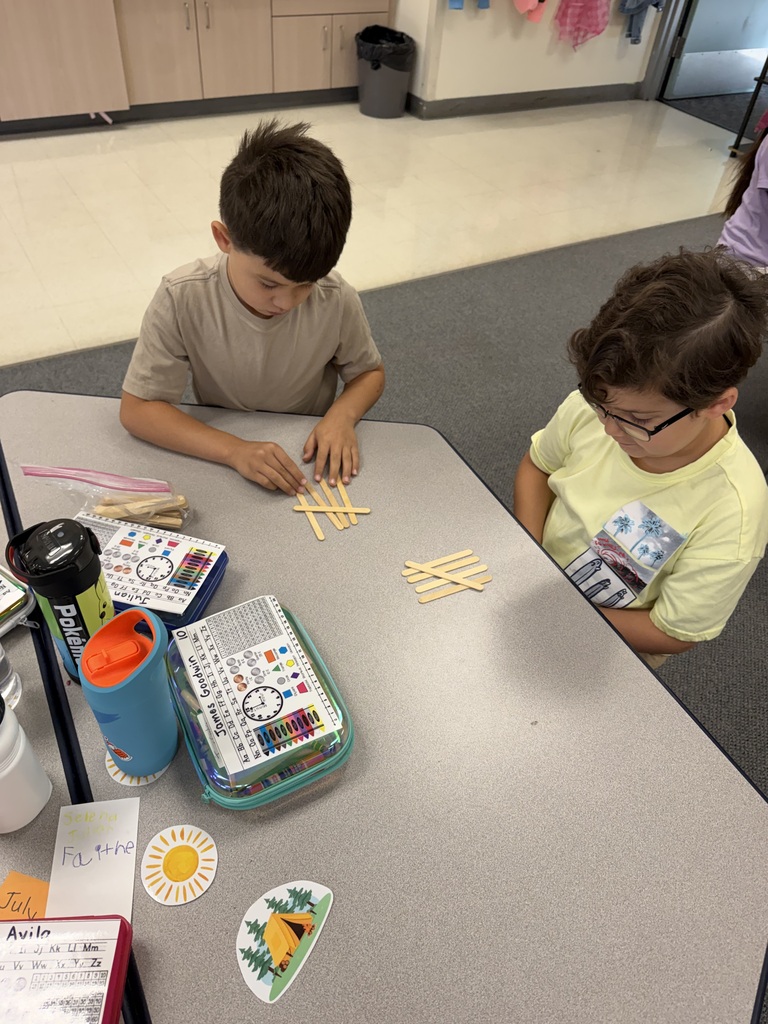 Two children seated at a table, engaged with wooden sticks, possibly participating in a learning activity.