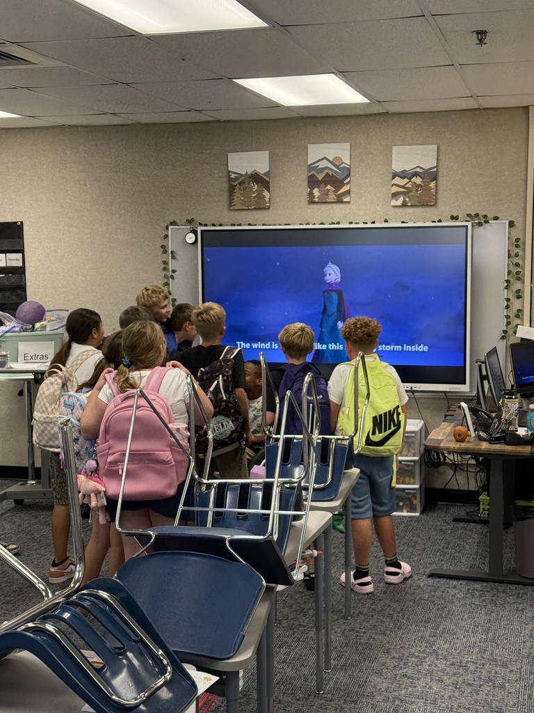Children stand around a large screen displaying a film. Most carry backpacks. A beige wall holds framed art.