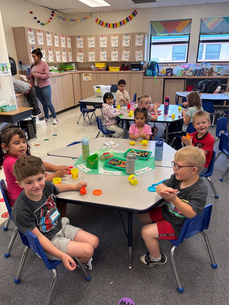 A classroom with children sitting at tables. One table has plastic eggs with toys inside. A woman stands behind them.