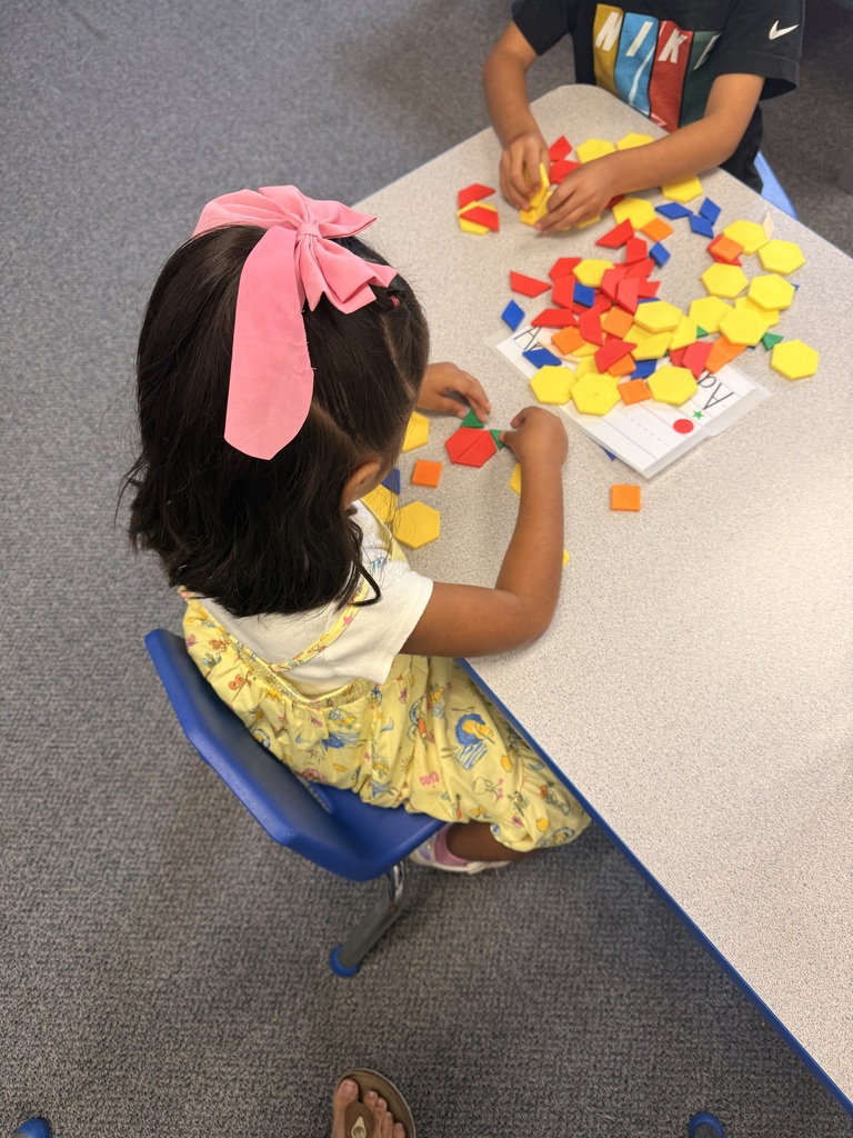 A child in a yellow dress sits at a table arranging colorful shapes, accompanied by another child nearby.