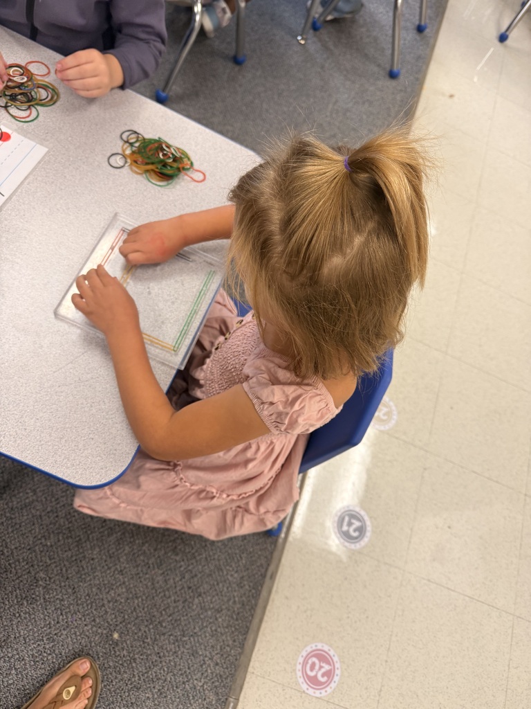 A young child in a pink dress sits at a table, working on a task. Various objects and papers are on the table.