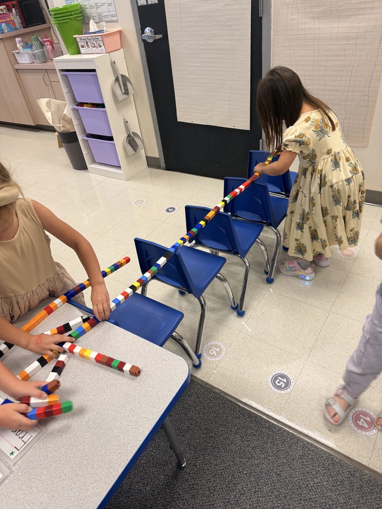 Two children wearing masks build colorful structures with plastic blocks at tables in a classroom.