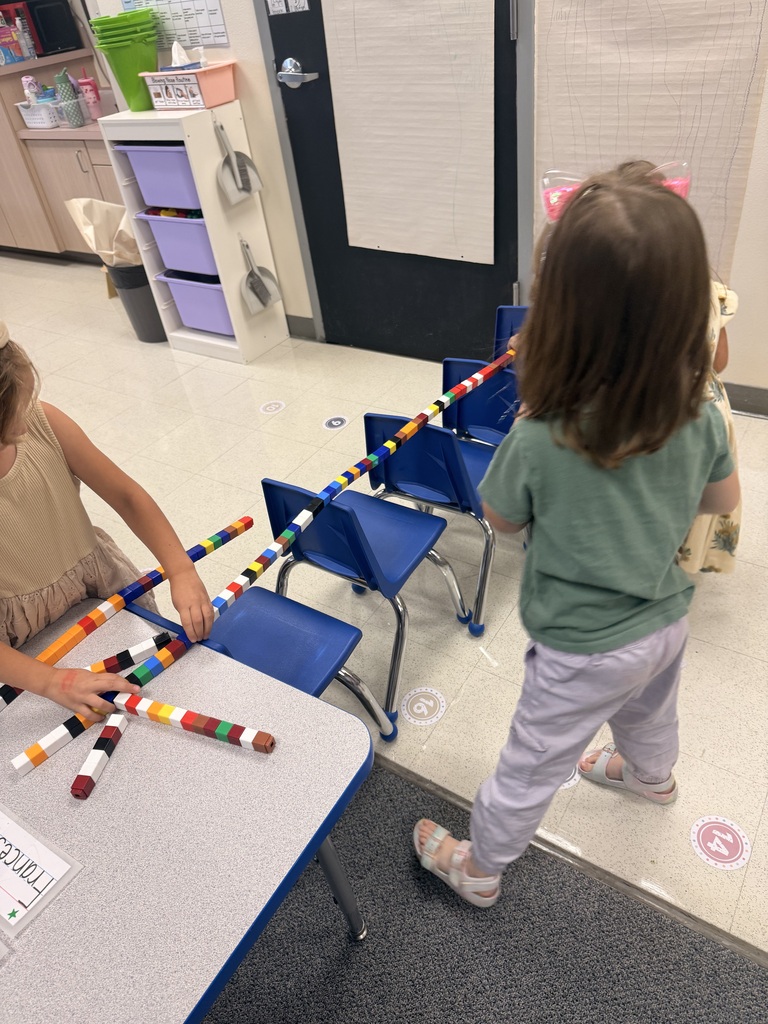 Two children play with colorful building blocks on a table, with chairs and a door in the background.