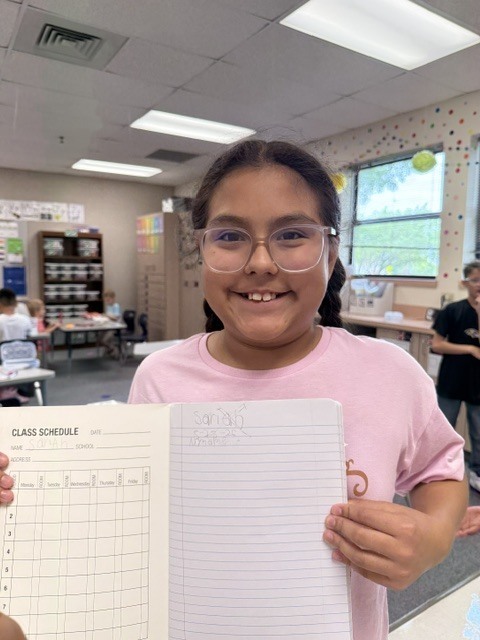 Child in a pink T-shirt holds open notebook displaying class schedule while smiling in a classroom.